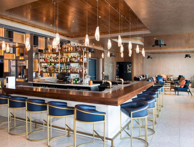 A stylish bar with modern seating, pendant lights, and a fully stocked back wall, accompanied by a cozy lounge area with armchairs.