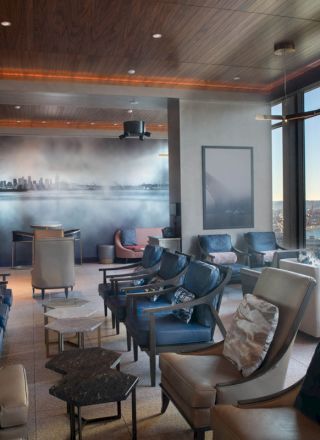 The image shows a modern lounge with plush seating, large windows offering a scenic view of the water, and a city mural on the wall.
