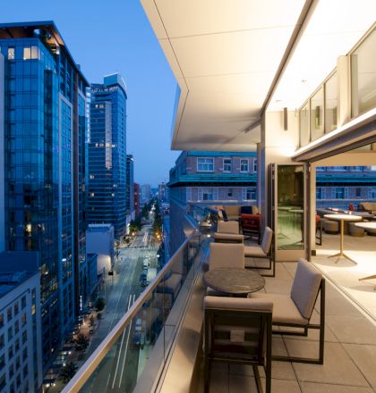 A modern rooftop terrace with seating overlooks a cityscape at dusk, featuring tall glass buildings and street lights below.