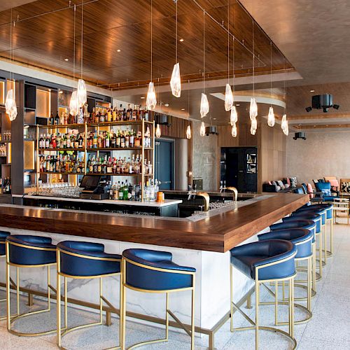 A stylish bar with a central counter, blue stools, hanging lights, and a variety of bottles on shelves behind the counter.