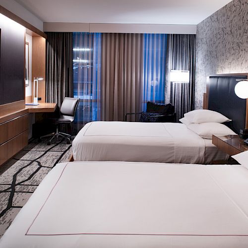 A modern hotel room with two beds, a TV, a desk with a chair, patterned wallpaper, and a carpet, illuminated by soft lights.
