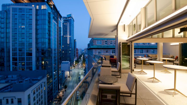 A modern urban balcony at dusk with cityscape views, featuring outdoor seating and tables under warm lighting.
