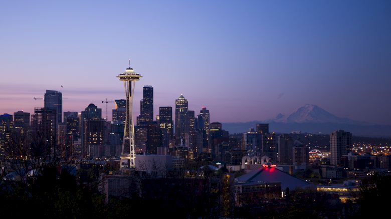 Skyline of a city at dusk with a distinctive tower in the forefront and a mountain in the background, lights illuminating buildings.