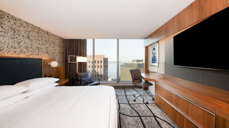 A modern hotel room with a large bed, desk, TV, and window view of city buildings and water, featuring stylish decor and wall art.