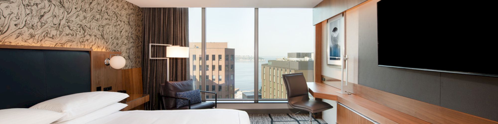 A modern hotel room with a large bed, wood furnishings, a desk, chair, and a flat-screen TV, overlooking city buildings through a large window.